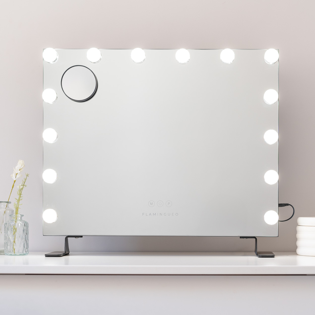 Un gran espejo de maquillaje con luces. Está sobre una superficie blanca, con un florero.