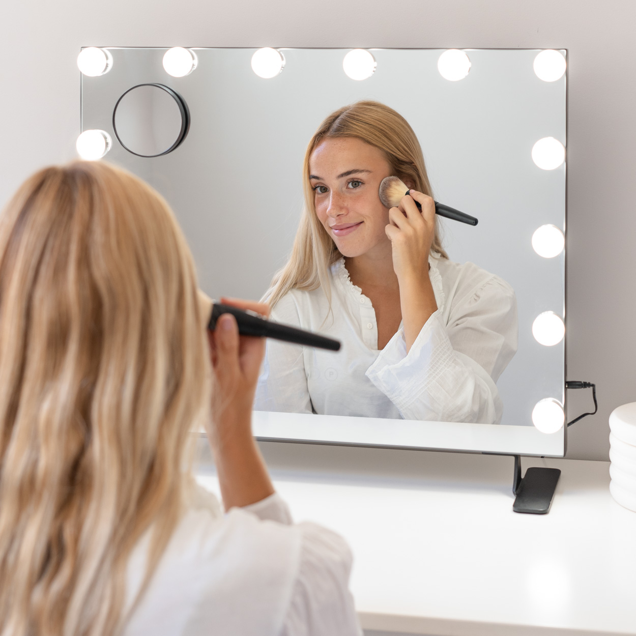 Una mujer maquillándose frente a un espejo de tocador con luces, sobre una mesa blanca con artículos de maquillaje.
