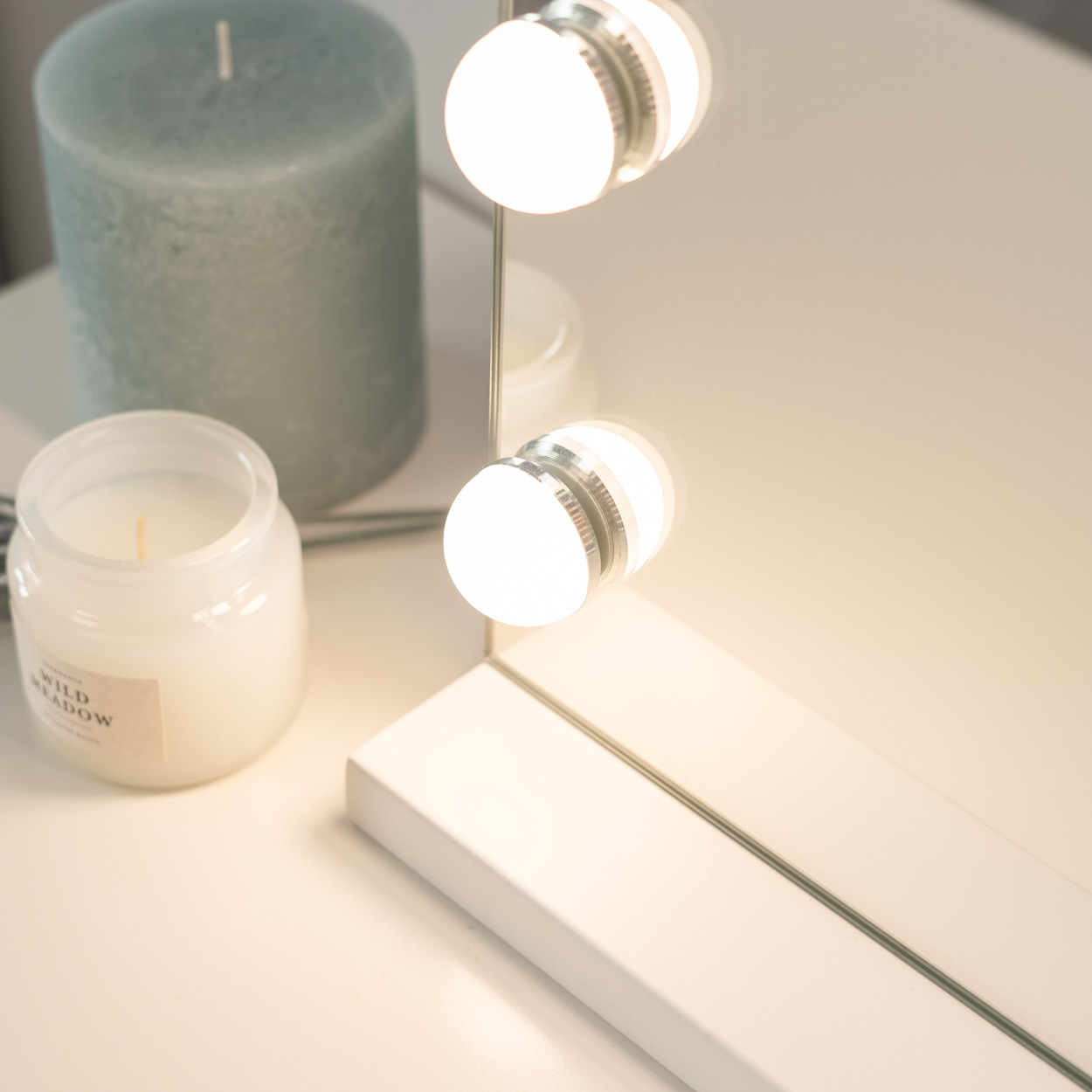 Espejo de maquillaje con luces sobre una mesa blanca. Velas visibles.