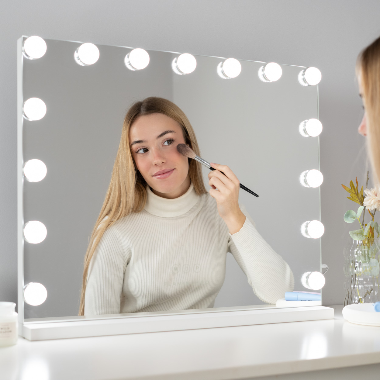 Mujer aplicándose maquillaje frente a un espejo con luces. Mesa y fondo blancos.