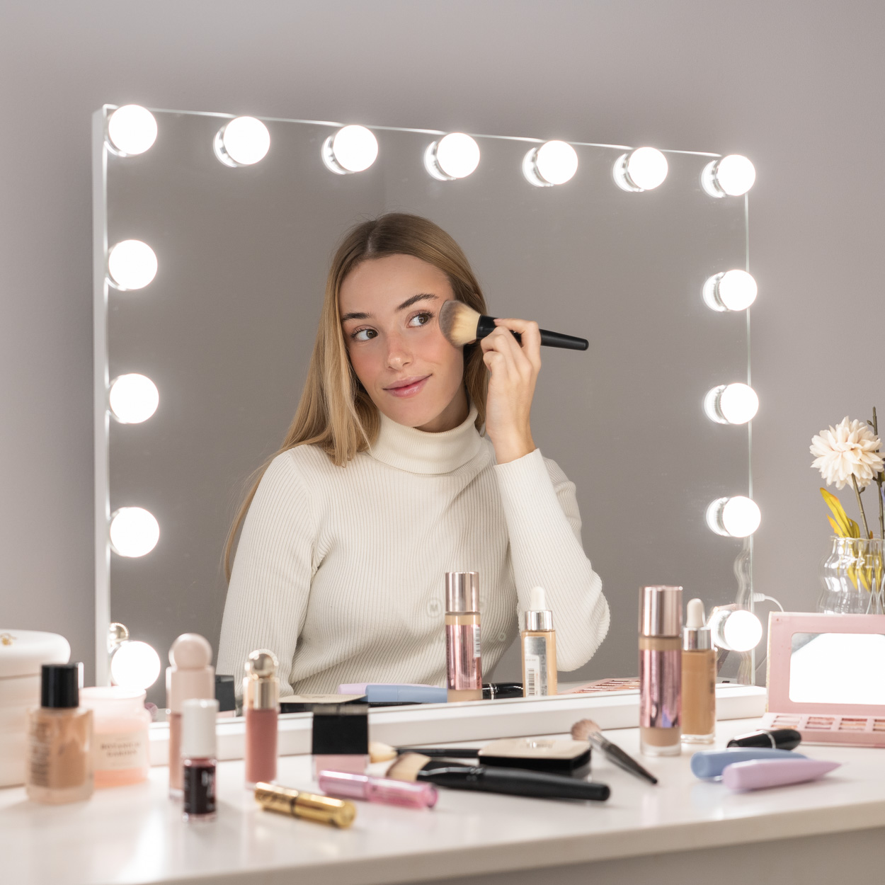 Mujer aplicándose maquillaje frente a un espejo iluminado. Productos de maquillaje en la mesa blanca. Fondo blanco.