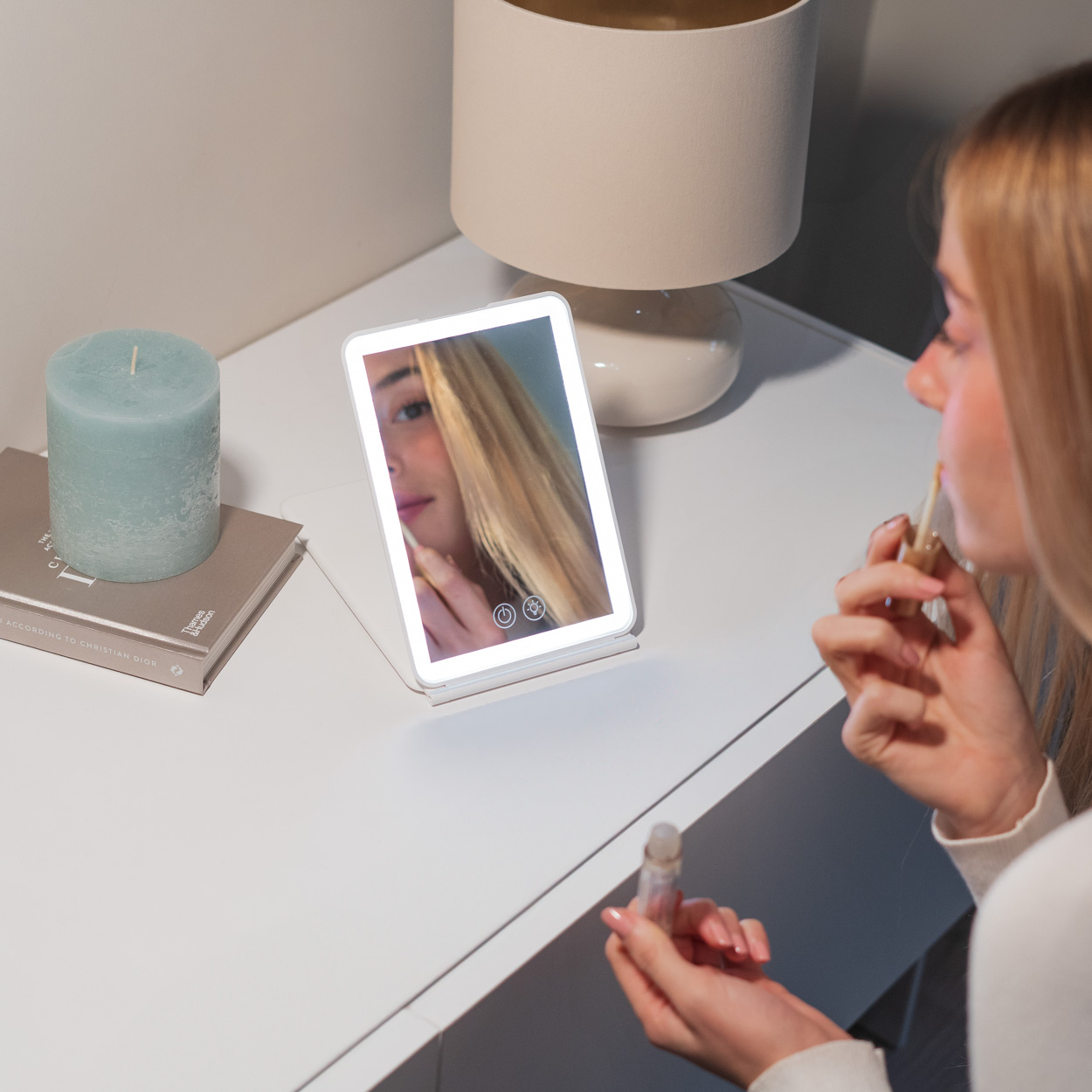 Mujer aplicándose lápiz labial frente a un espejo de tocador iluminado. Vela y lámpara sobre la mesa.