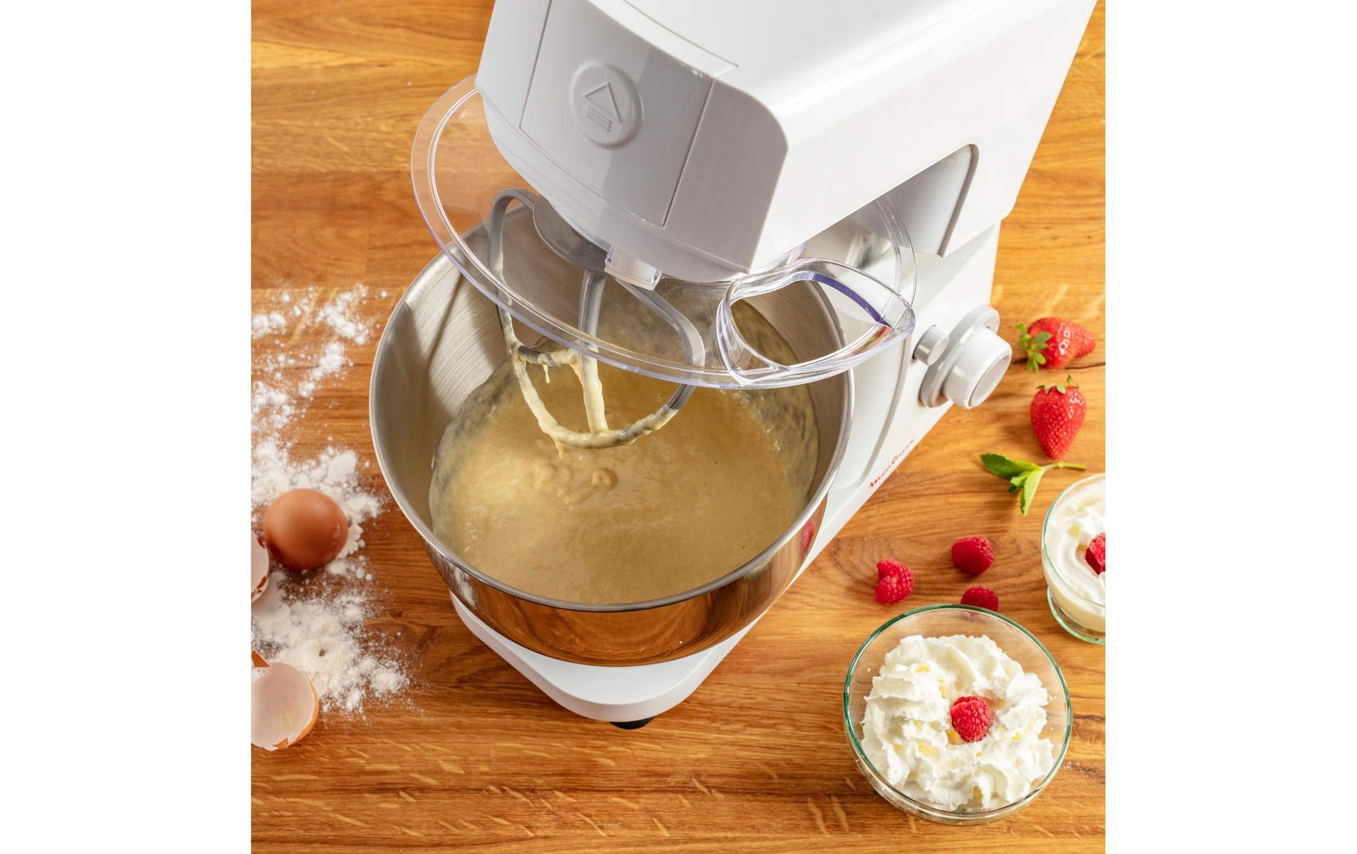 Un mixer bianco con una ciotola, pastella all'interno e ingredienti nelle vicinanze su una superficie di legno.