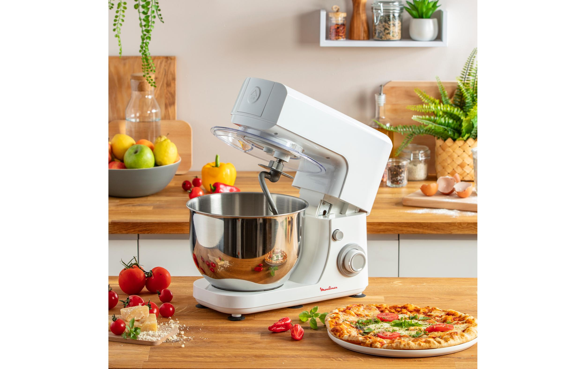 Un mixer da cucina bianco con una ciotola in acciaio inox e accessori su un piano di lavoro in legno. Gli ingredienti sono nelle vicinanze.