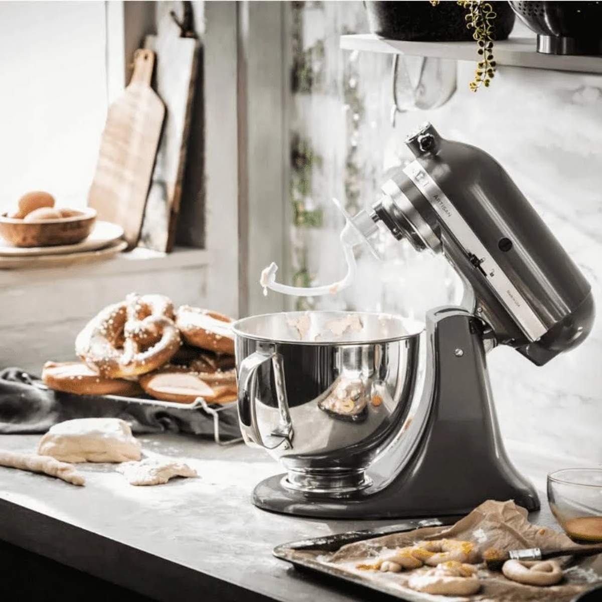 Gri KitchenAid mikser hamuru karıştırıyor. Hamur ve simitler pencerenin yanındaki tezgahta.