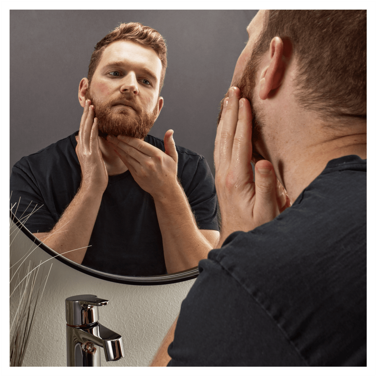 Un hombre se mira en un espejo, aplicándose producto en la barba. El espejo es redondo, el fondo es gris.