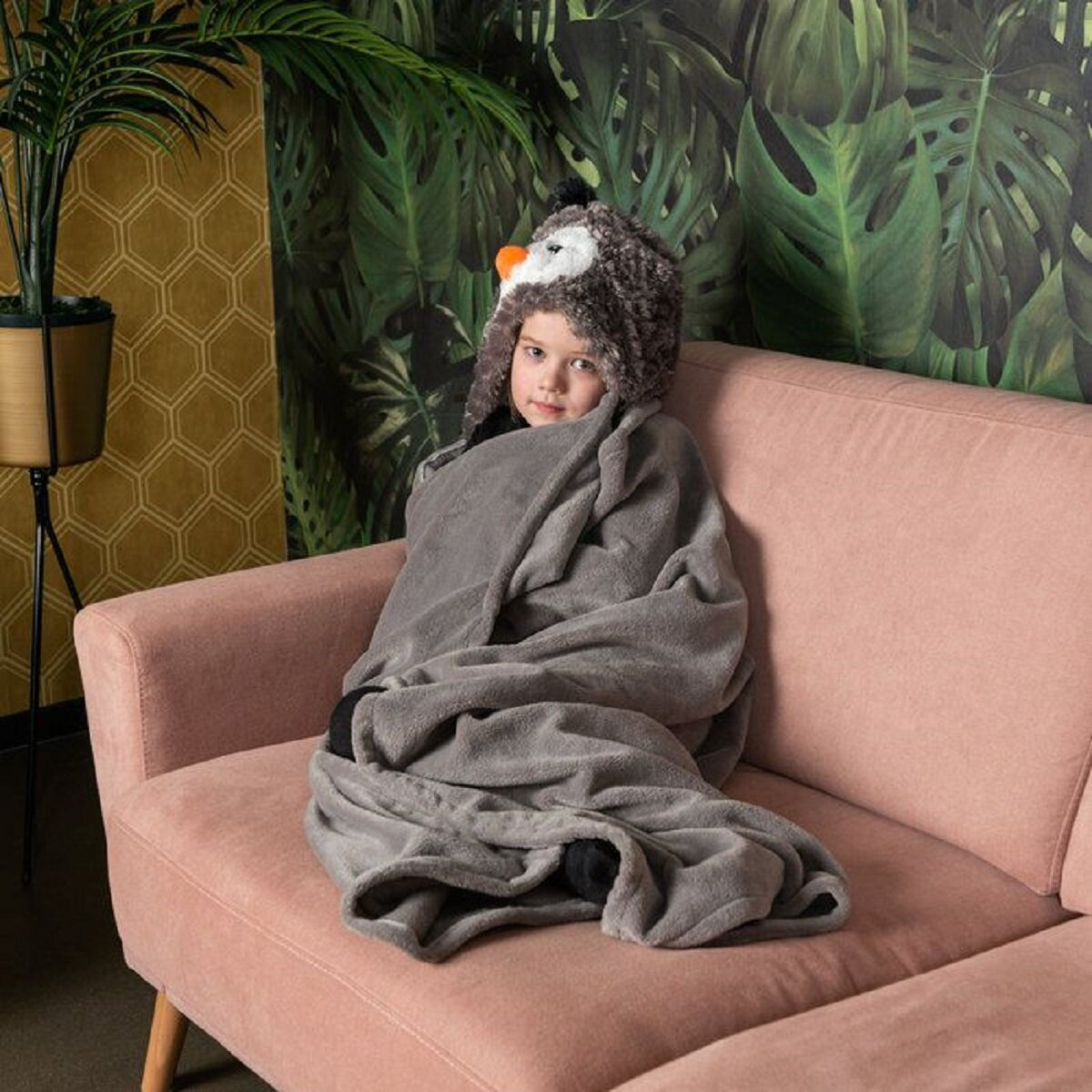Ein Kind mit Pinguinhut, eingewickelt in eine graue Decke, sitzt auf einem rosa Sofa, grüne Pflanze im Hintergrund.