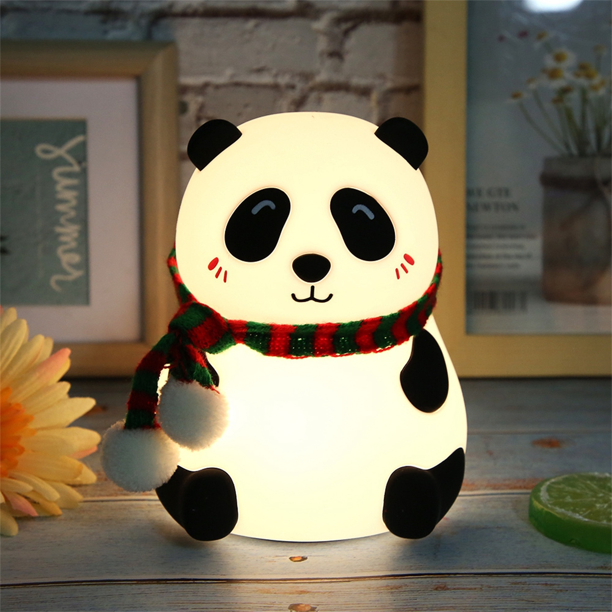 Panda-förmiges Nachtlicht mit rotem und grünem Schal. Es leuchtet und sitzt auf einer Holzoberfläche.
