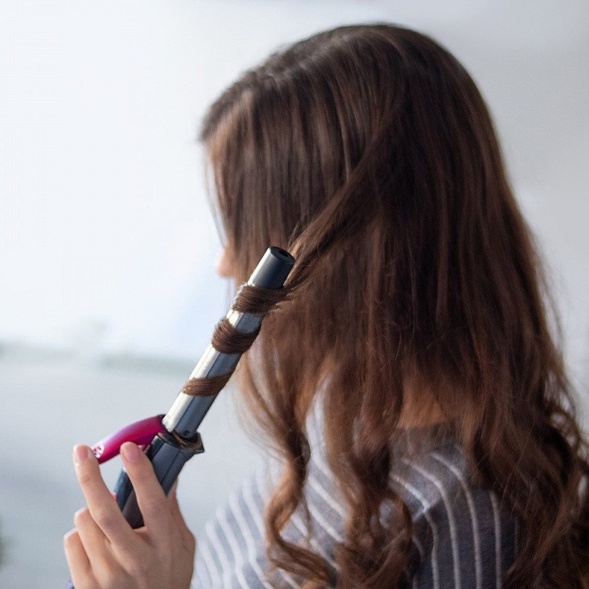 Una mujer se riza el pelo con un rizador. El fondo es una pared blanca borrosa.