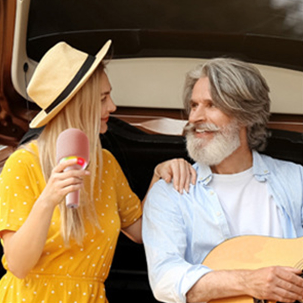 Ein Mann und eine Frau stehen in der Nähe eines Autos. Die Frau hält ein rosa Mikrofon, der Mann spielt Gitarre.
