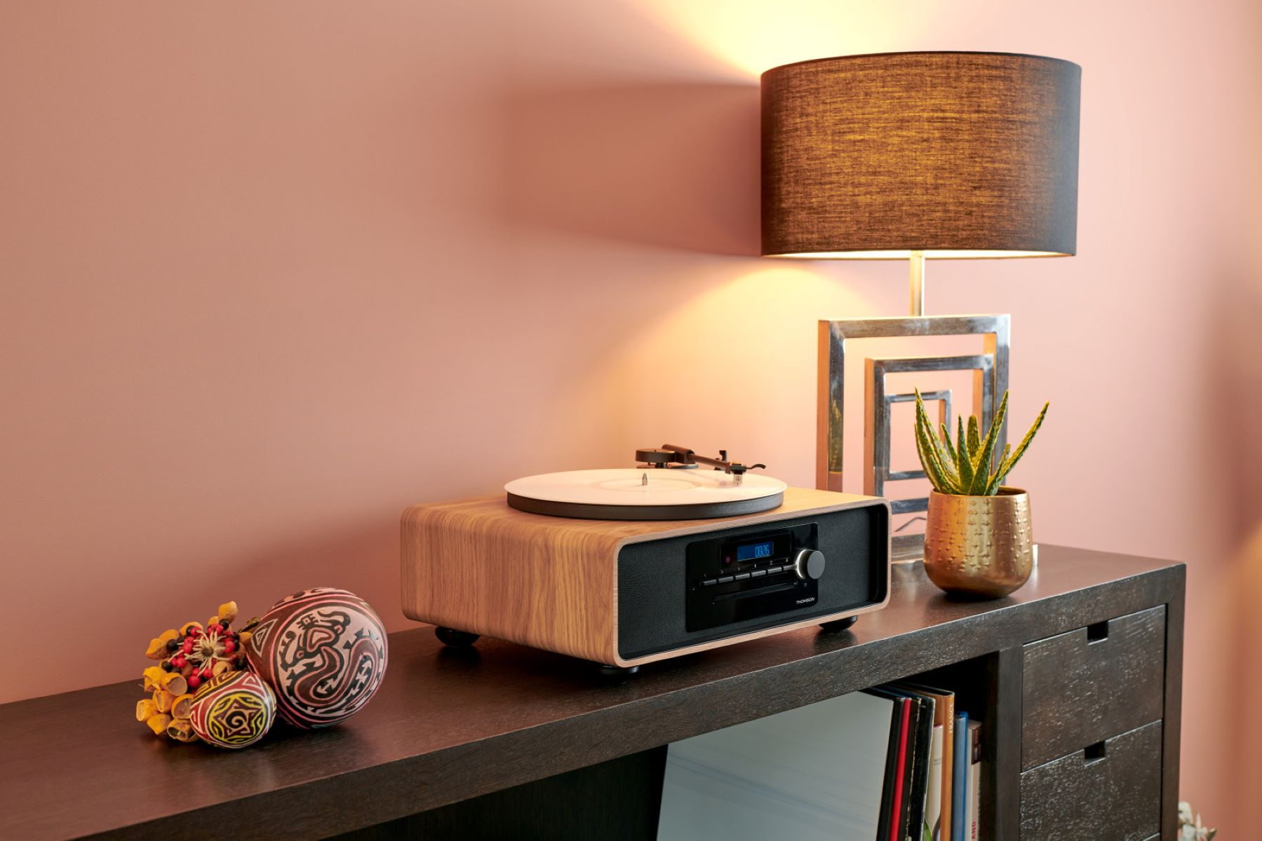 Drewniany gramofon na ciemnej szafce z lampą, dekoracjami i rośliną w tle.