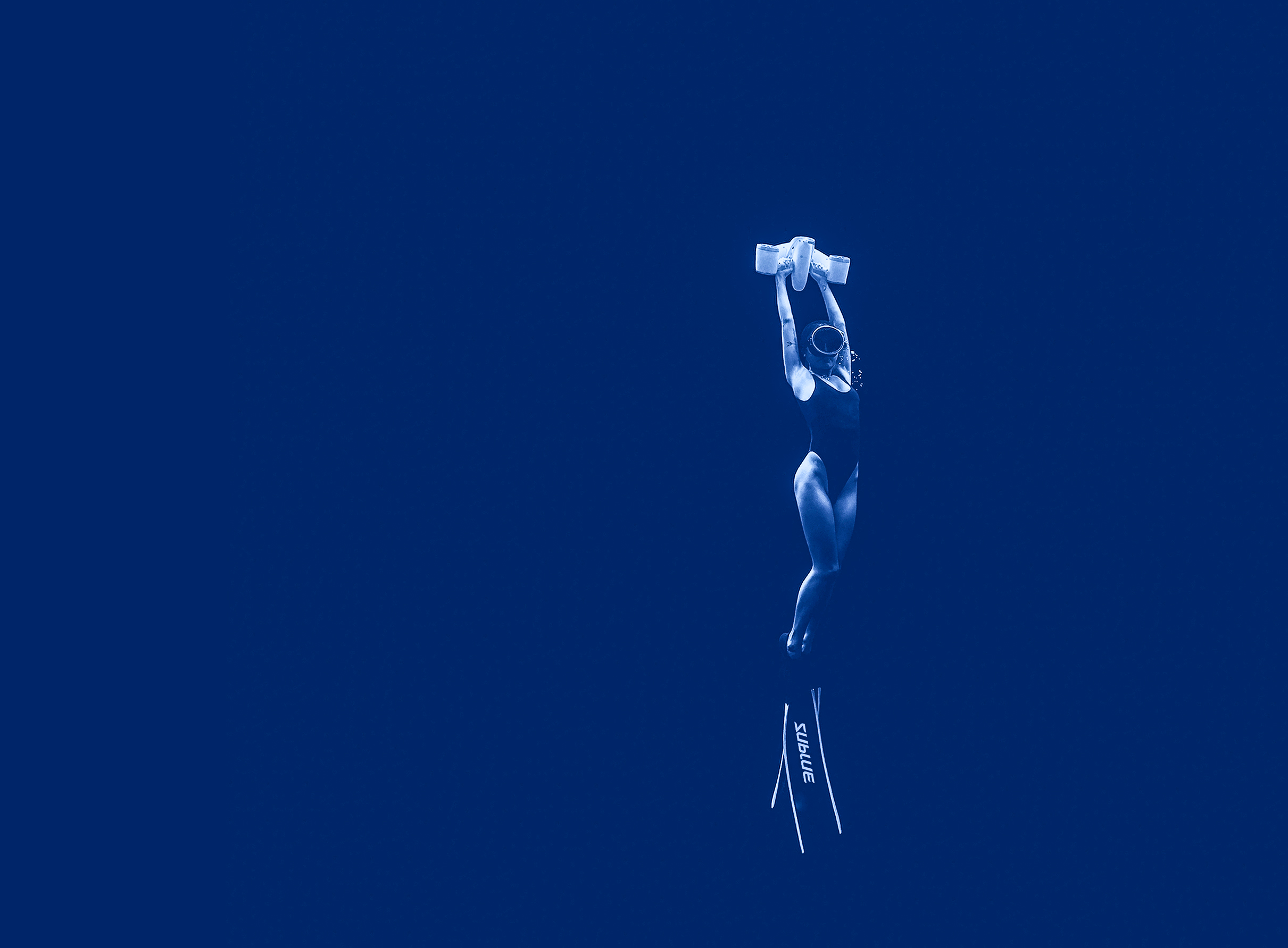 A woman in a black swimsuit is underwater, with fins, arms up, in blue water.