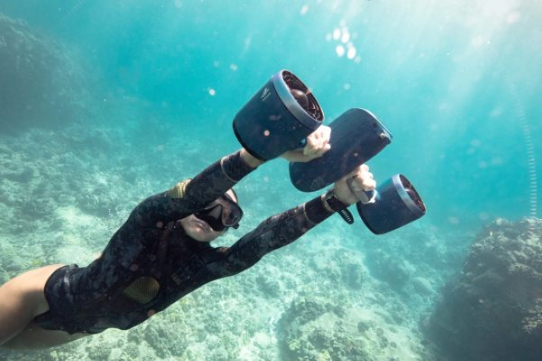 A person with a mask, holding two underwater scooters. They are underwater in the ocean.