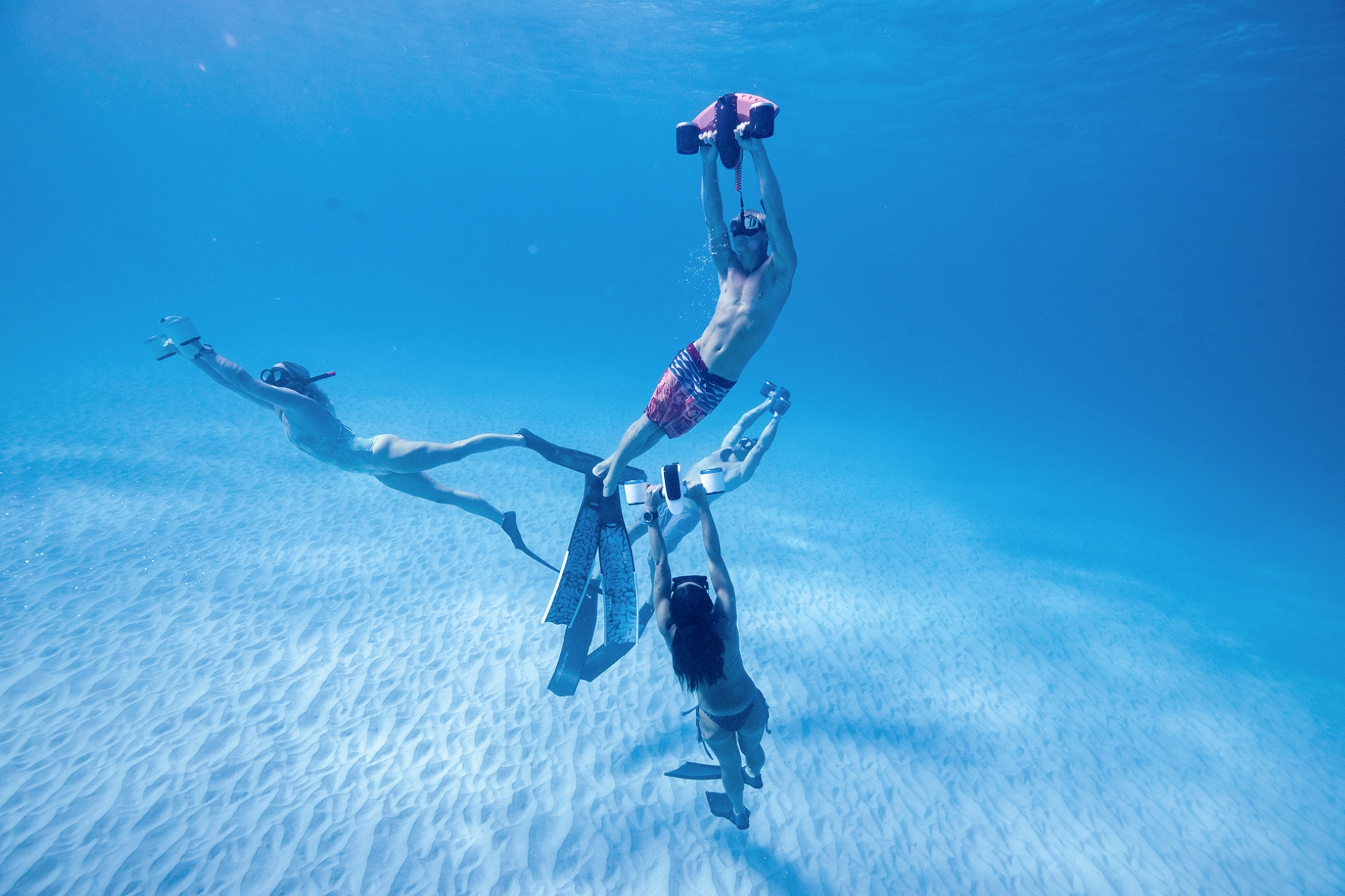 Underwater shot of four people, two with fins, playing with boards, in a blue ocean.