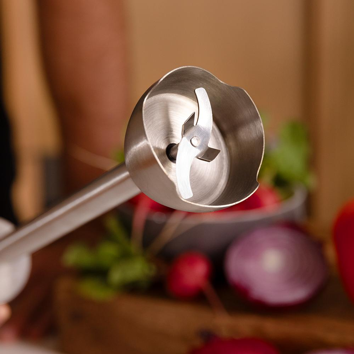 Głowica blendera ręcznego z ostrzami, zbliżenie, z rozmytym tłem warzyw, czerwonej cebuli i pomidorów.