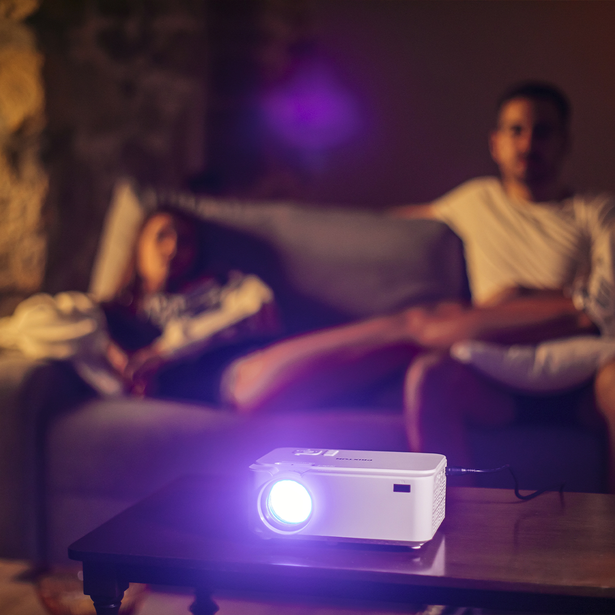 Projektor na stole rzuca światło. Dwie osoby siedzą na kanapie w tle.
