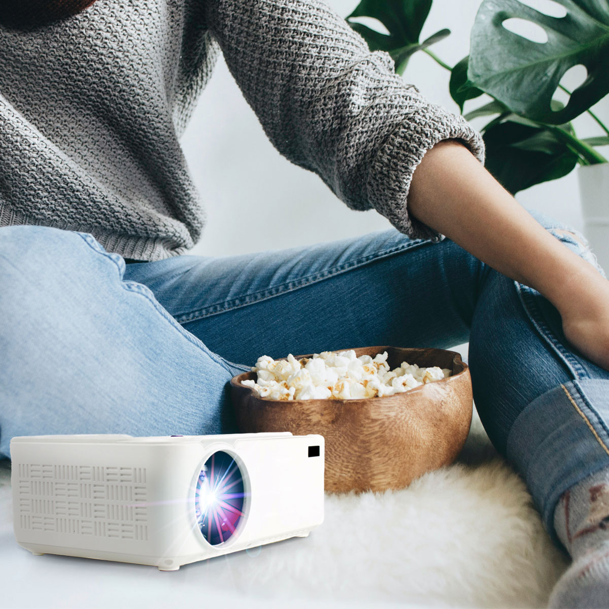 Osoba siedząca z projektorem i popcornem, przed zieloną rośliną.