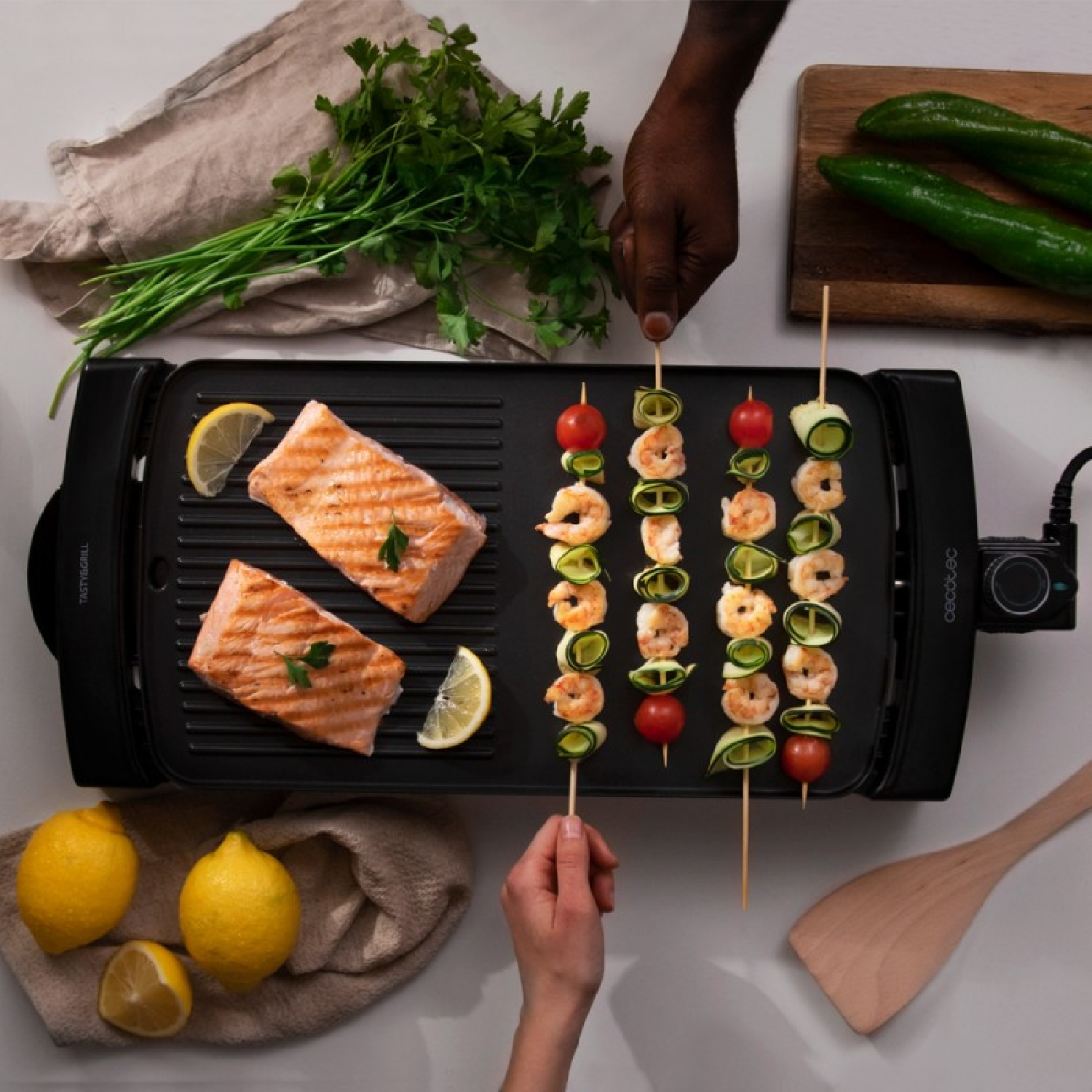 Parrilla con salmón, brochetas de camarones, limones, perejil y pepino. Una persona está asando.