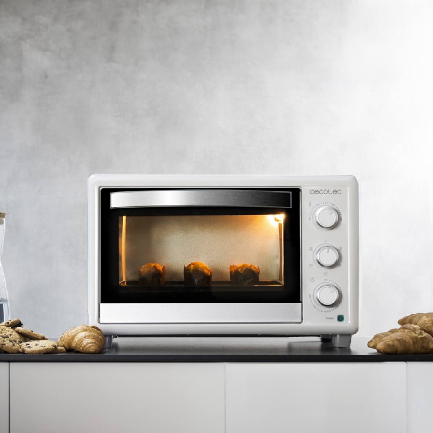 Een witte oven met drie muffins die erin bakken. De oven staat op een wit aanrecht.