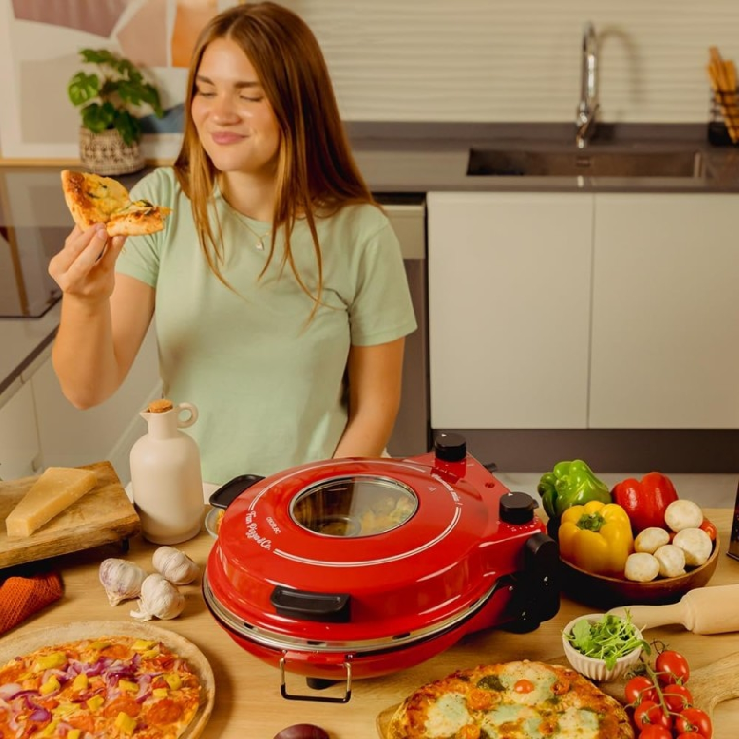 Una donna tiene in mano una fetta di pizza vicino a un forno per pizza rosso, con ingredienti come aglio e peperoni sul tavolo.