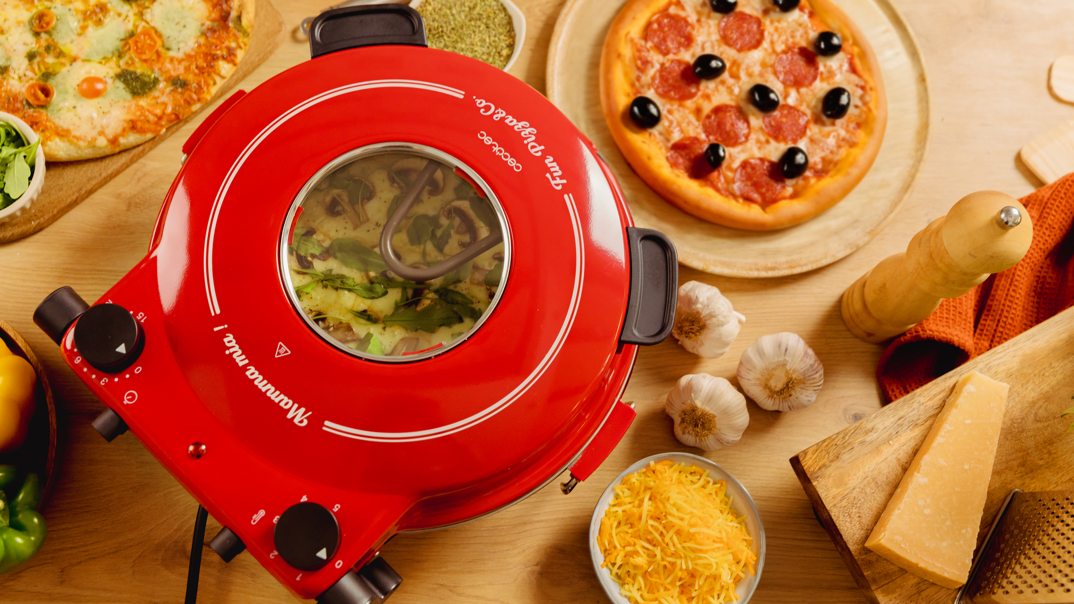 Un forno per pizza rosso con finestra in vetro. Pizze e ingredienti sulla superficie di legno intorno al forno.
