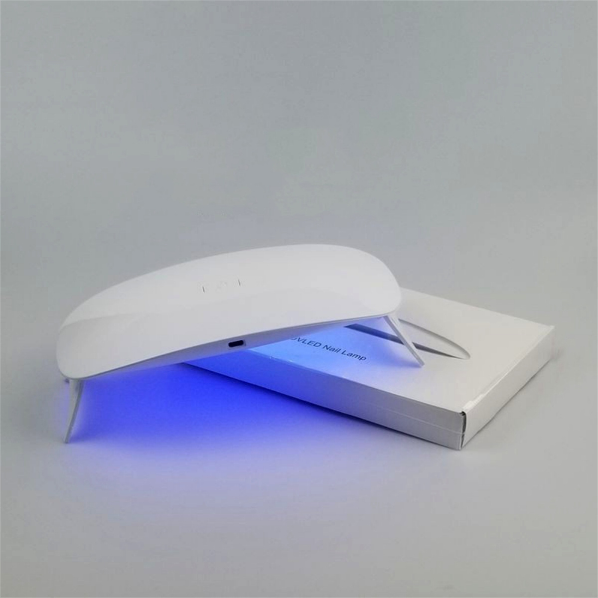 Eine weiße UV-Nagellampe mit blauem Licht im Inneren und eine weiße Box. Die Lampe steht auf einem hellgrauen Hintergrund.