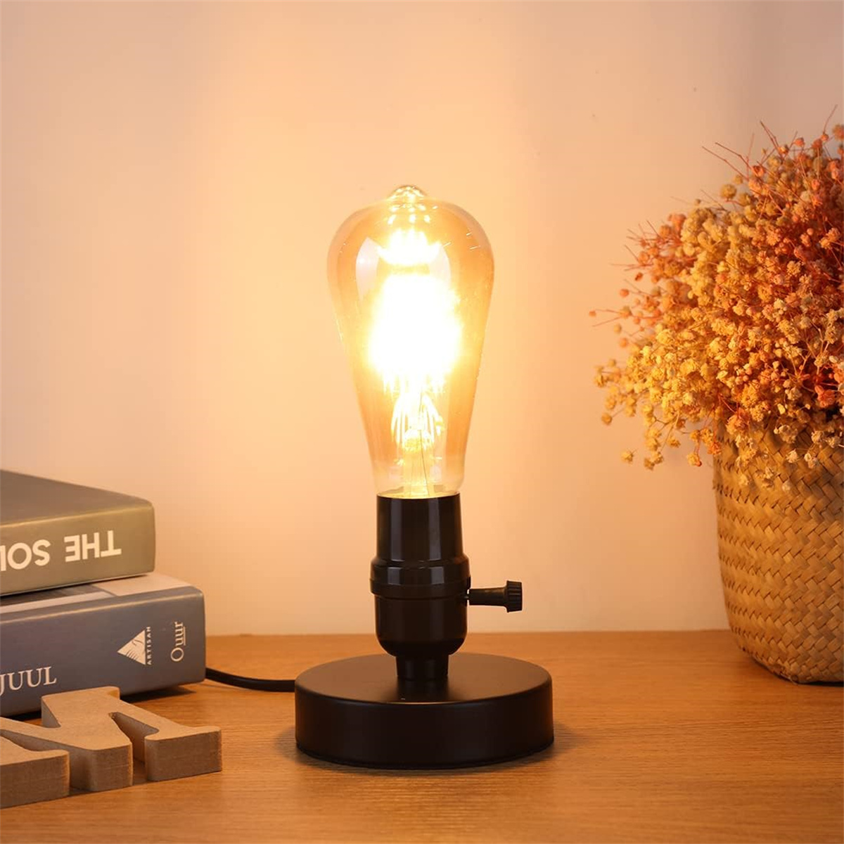 Eine Lampe mit Glühbirne im Vintage-Stil auf einem Tisch, Bücher und Blumen im Hintergrund.