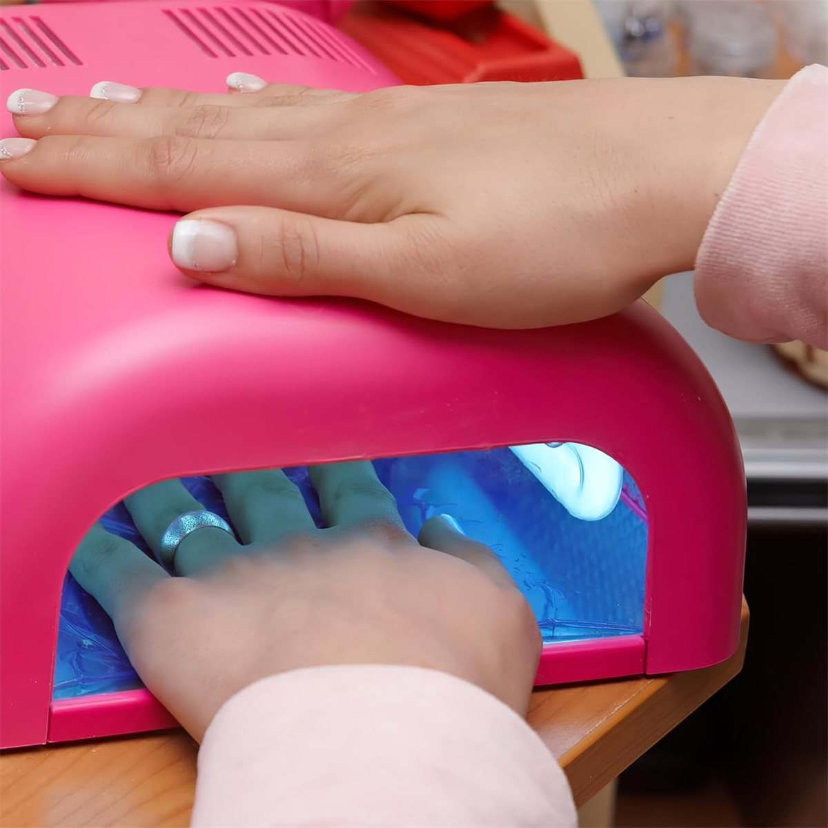 Hände in einem pinkfarbenen Nageltrockner mit blauem Licht, eine Hand mit Ring.
