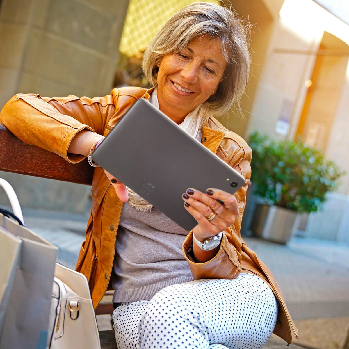 Una mujer sentada en un banco, sosteniendo una tableta y sonriendo. Lleva una chaqueta de cuero y pantalones blancos con lunares.