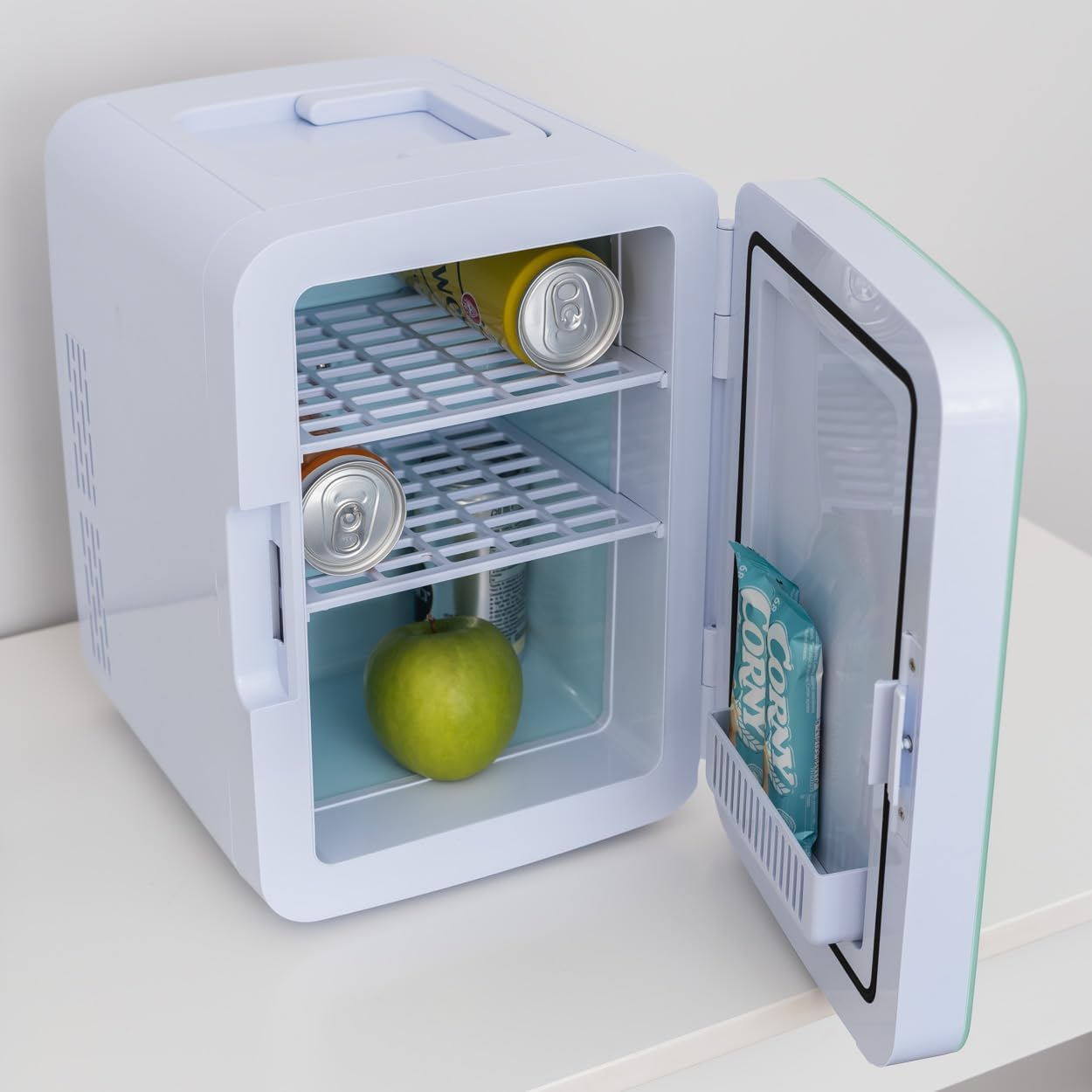 Mini-nevera blanca abierta, que contiene latas, una manzana y un aperitivo.