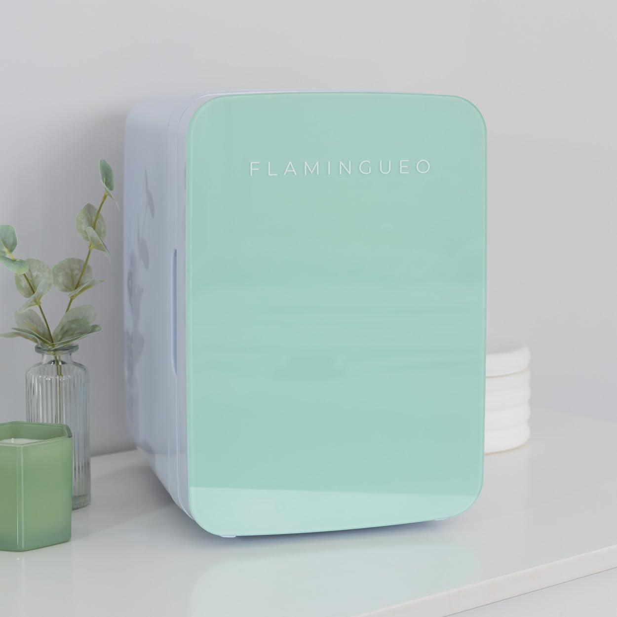 Una mini-nevera verde menta con borde blanco, etiquetada FLAMINGUEO. Un jarrón, vela y planta cerca.