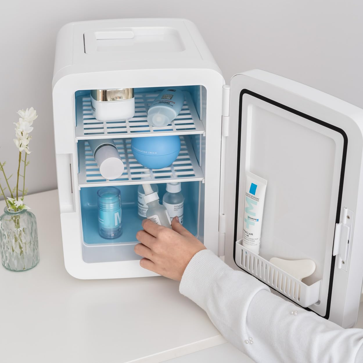 Un mini-refrigerador blanco abierto, revelando cosméticos. Una mano se mete. Un florero a la izquierda.