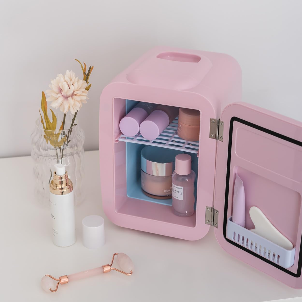 Una mini-nevera rosa con cosméticos en su interior, junto a un rodillo de cuarzo rosa y un jarrón.
