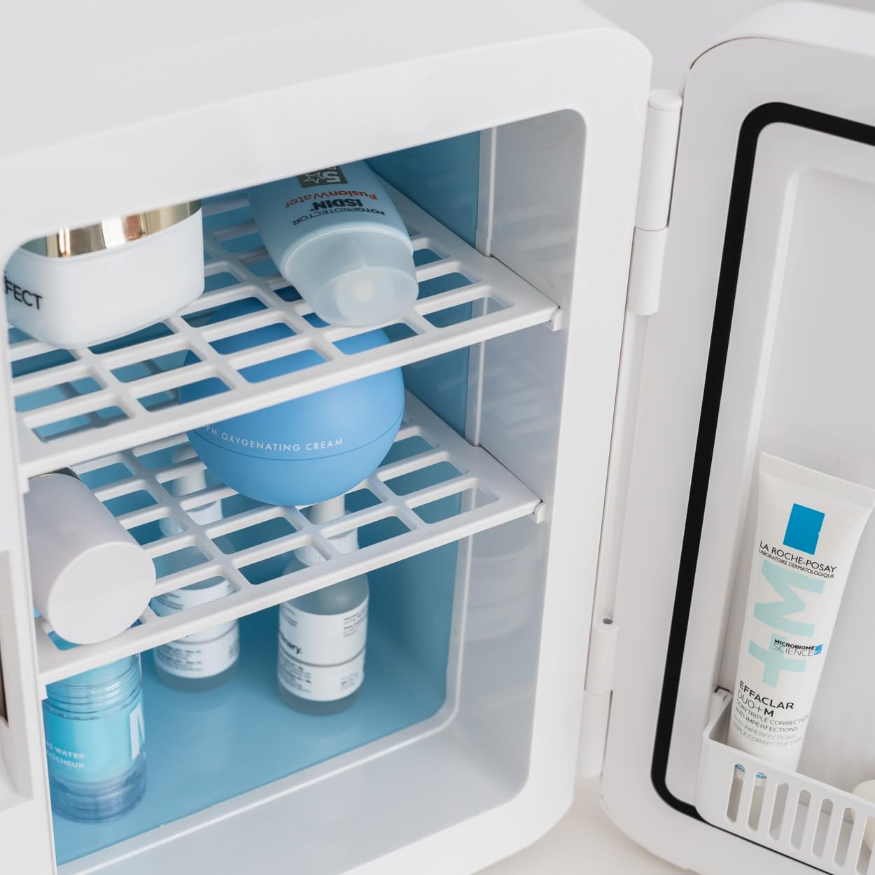 Mini-nevera blanca abierta con productos para el cuidado de la piel. Interior azul y estantes blancos.