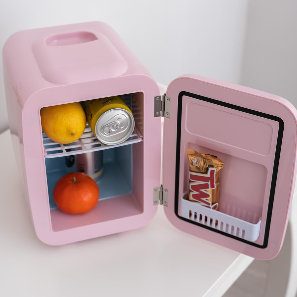 Min nevera rosa con puerta abierta, con un limón, una lata, una mandarina y una barra de caramelo.