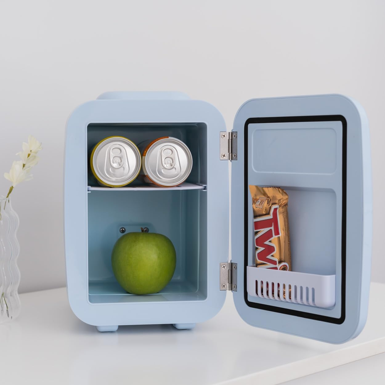 Pequeño frigorífico azul con puerta abierta. Dentro: latas y barra Twix.