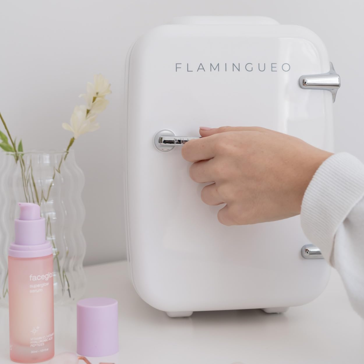 Una mini nevera blanca con una mano abriendo la puerta. Hay cosméticos y un jarrón cerca.