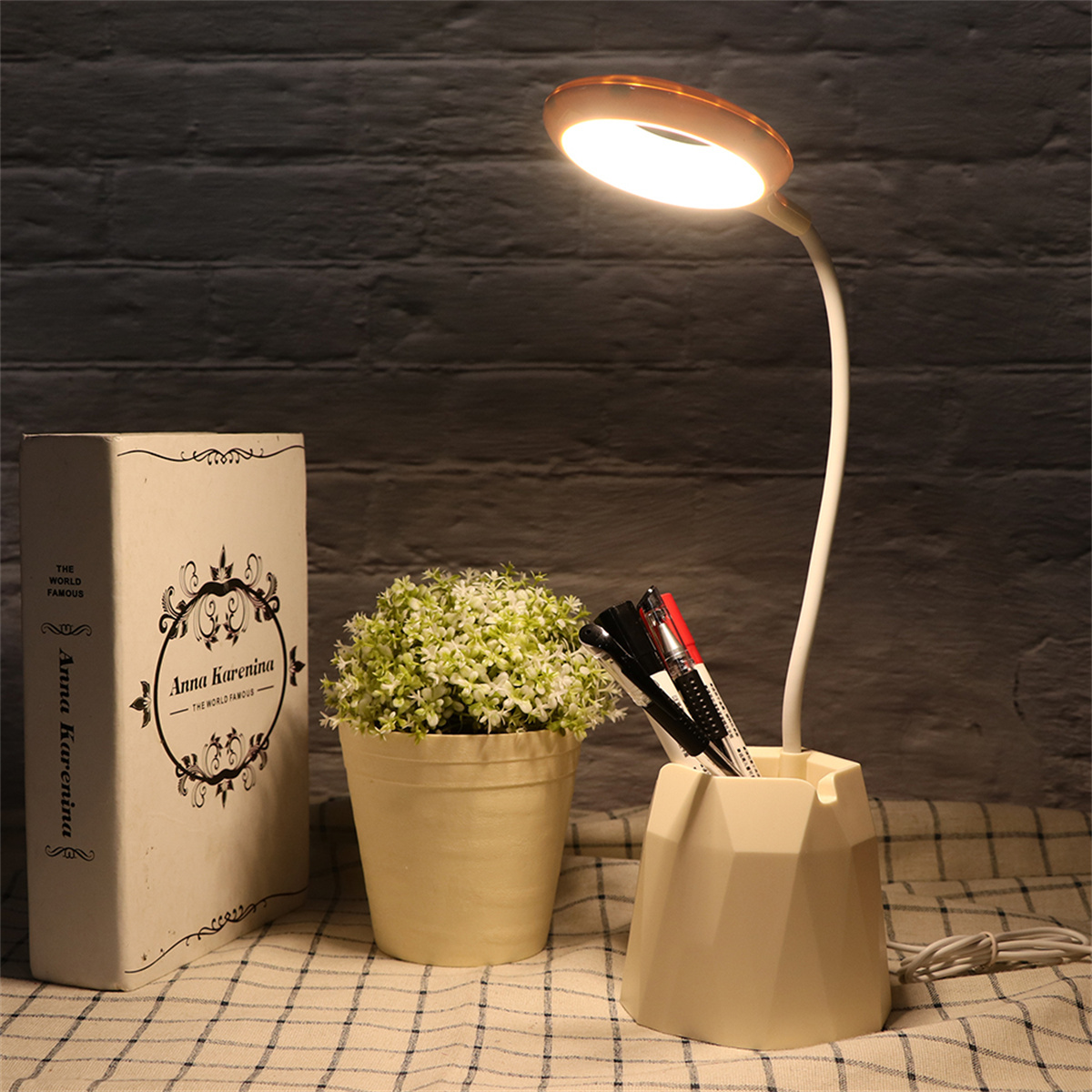 Eine Schreibtischlampe, eine Topfpflanze, ein Buch und Stifte in einem Stifthalter auf kariertem Tuch.