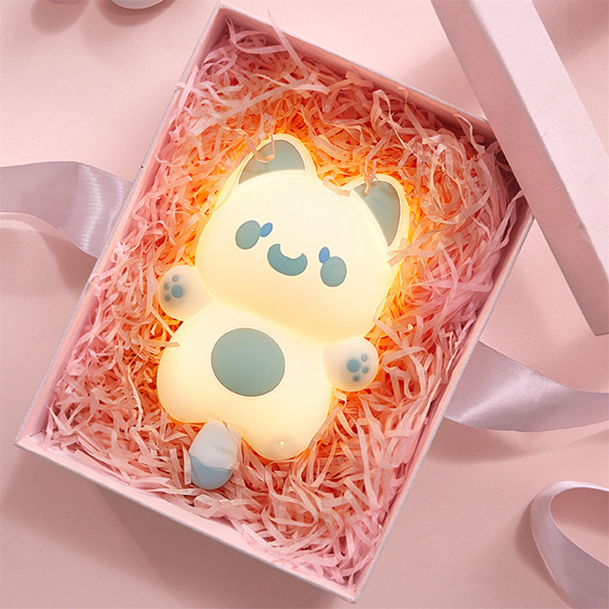 Ein katzenförmiges Nachtlicht liegt in einer rosa Geschenkbox mit geschreddertem Papier. Das Licht ist gelb mit blauen Akzenten.
