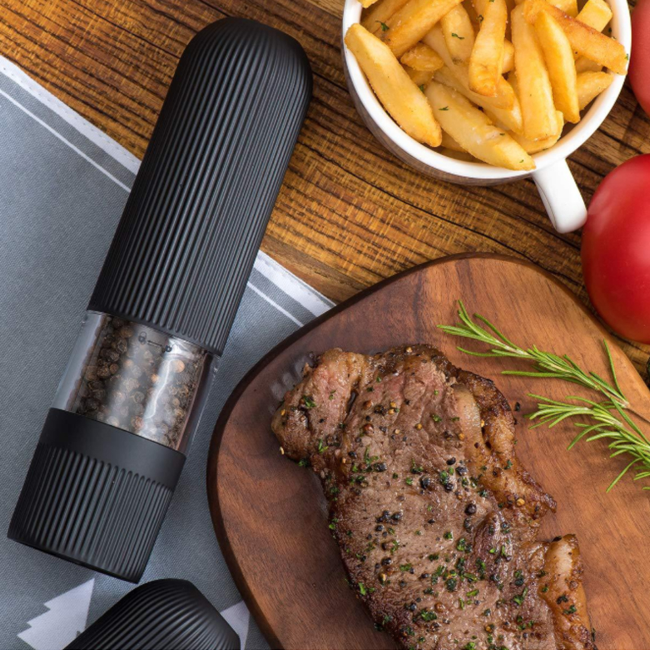 Eine schwarze Pfeffermühle neben Steak auf einem Holzbrett, Pommes und Tomate an der Seite.