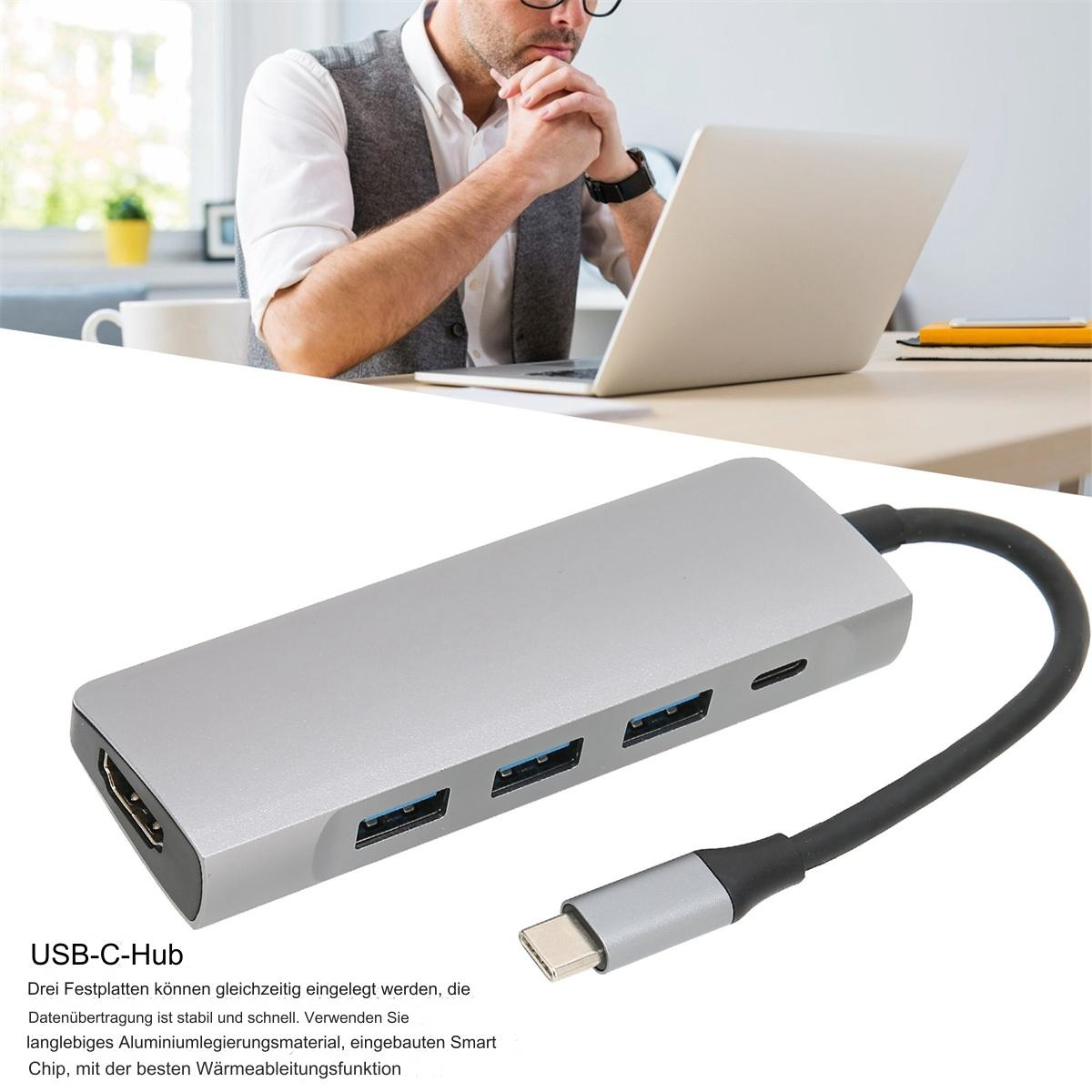 USB-C-Hub mit HDMI, USB-Anschlüssen. Person arbeitet im Hintergrund am Laptop.