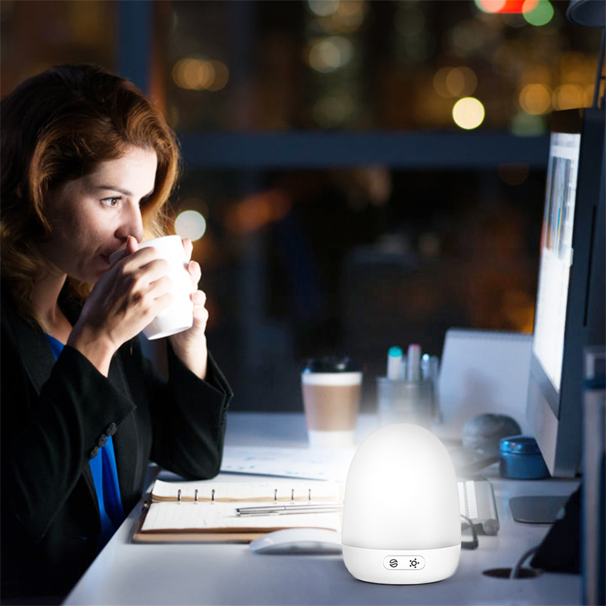 Eine Frau trinkt nachts Kaffee. Eine weiße Lampe beleuchtet den Schreibtisch. Computermonitor rechts.