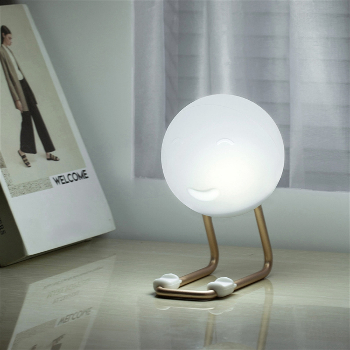 Weiße, lächelnde Mondlampe mit goldenen Beinen, steht auf einem Tisch. Buch und Fenster im Hintergrund.