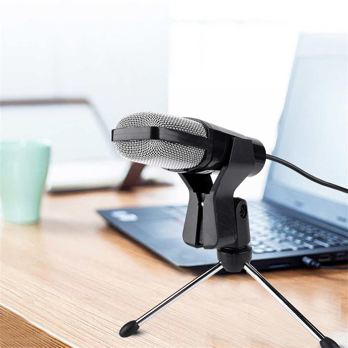 Ein schwarzes Mikrofon auf einem Stativ steht auf einem Holztisch mit Laptop und grüner Tasse im Hintergrund.