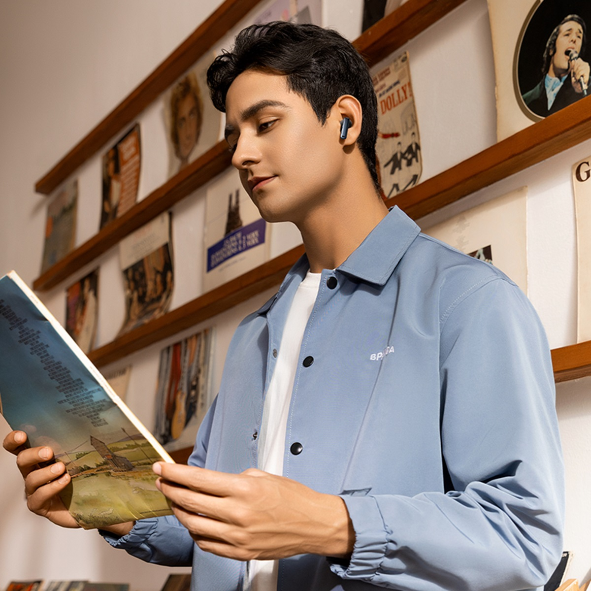 Młody mężczyzna w słuchawkach, czytający książkę. Jest przed ścianą płyt winylowych.