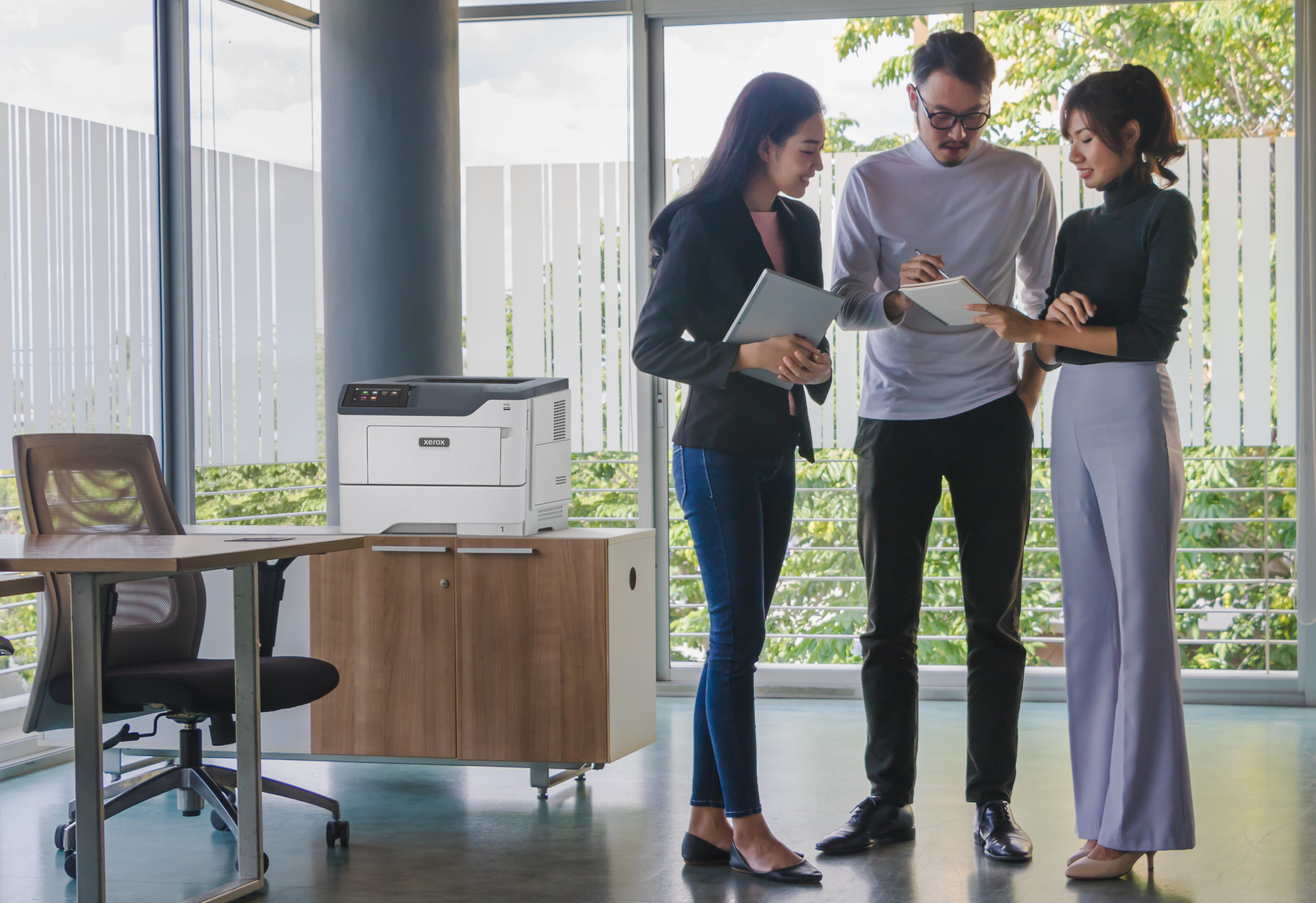 Scena biurowa z trzema osobami przy biurku i drukarką na szafce.