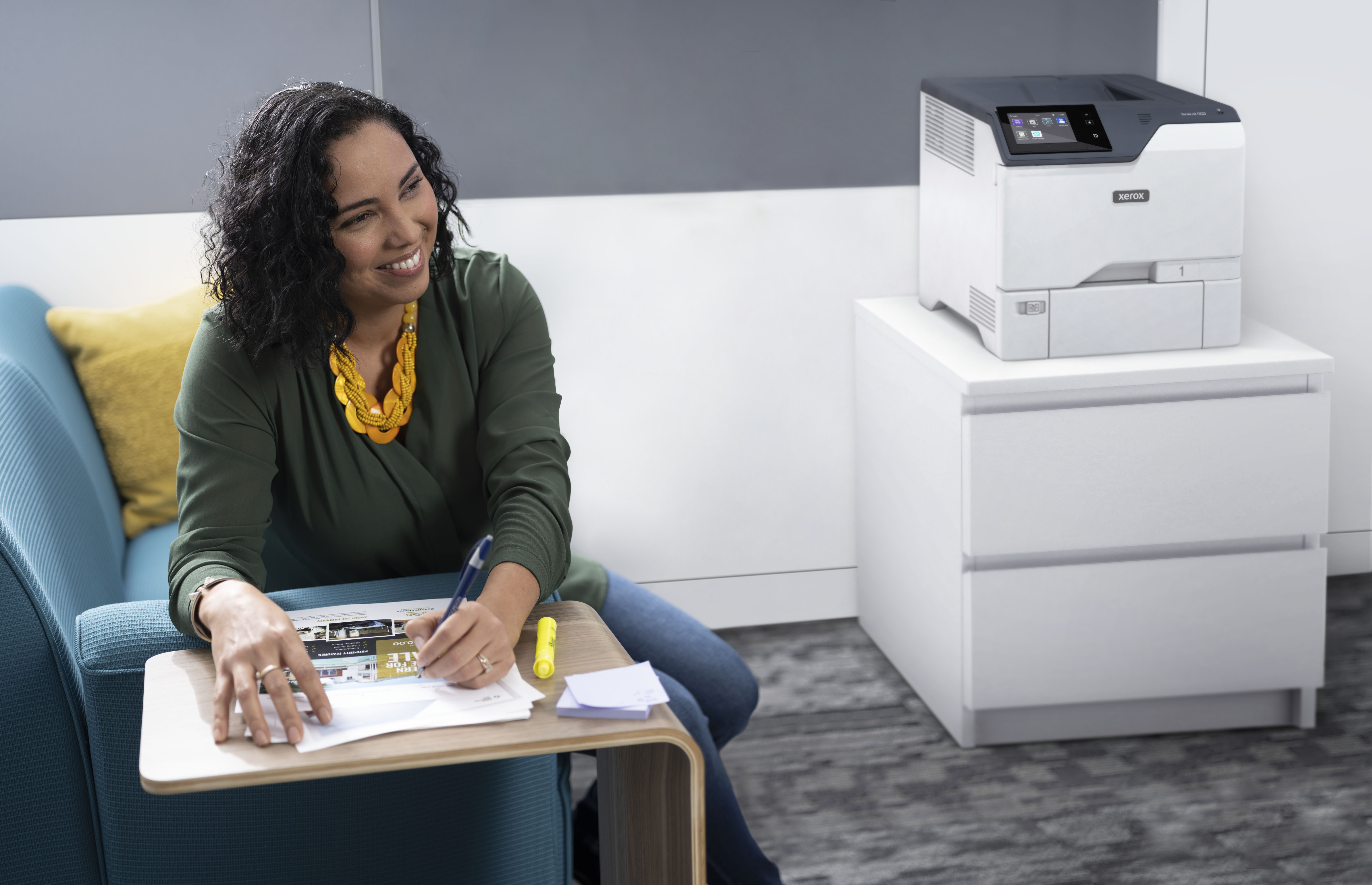 Kobieta siedzi na niebieskiej kanapie, pisząc obok białej drukarki na szafce.