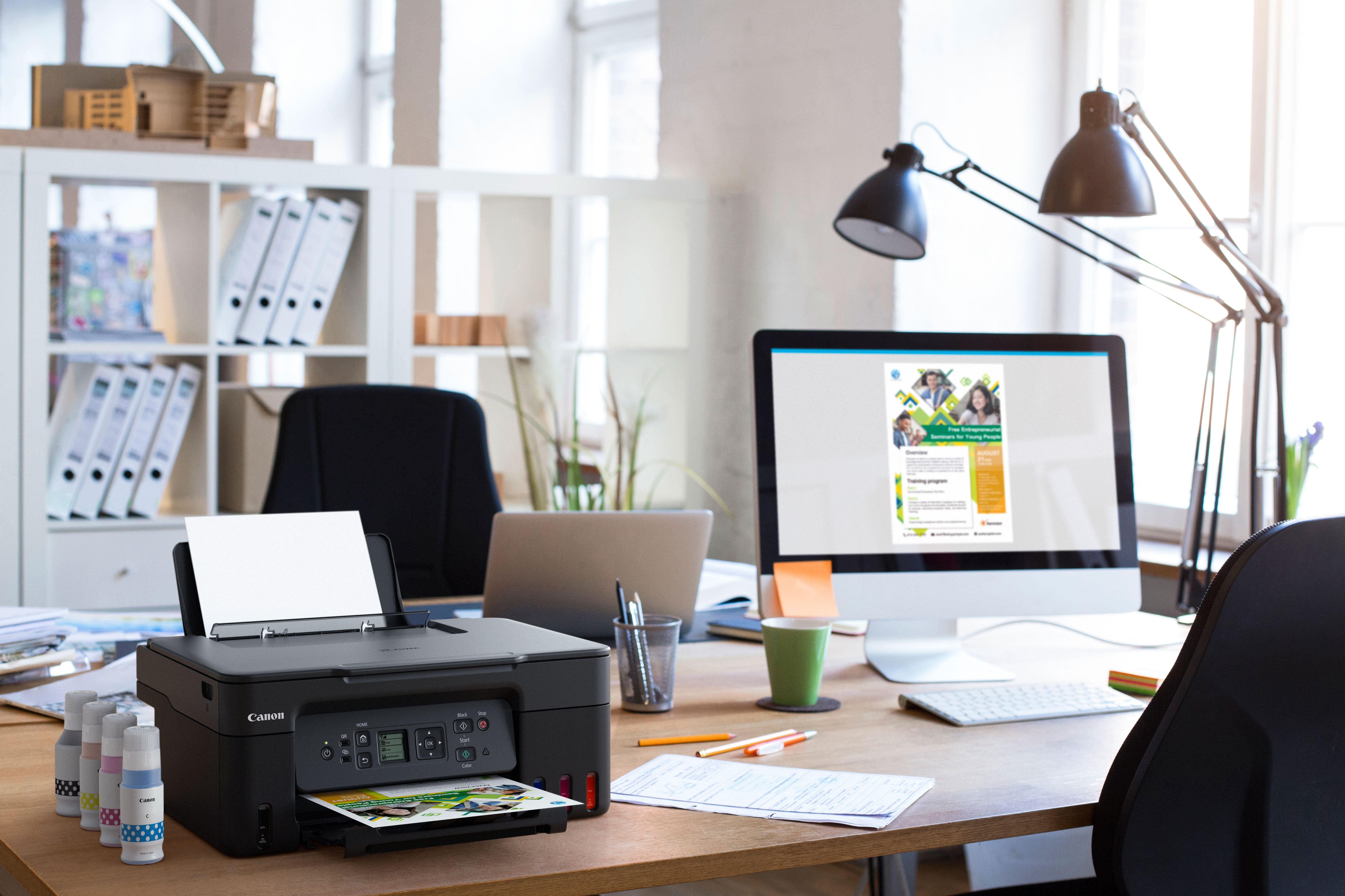 Biurko z drukarką, monitorem i butelkami z tuszem. Drukarka drukuje zdjęcie.