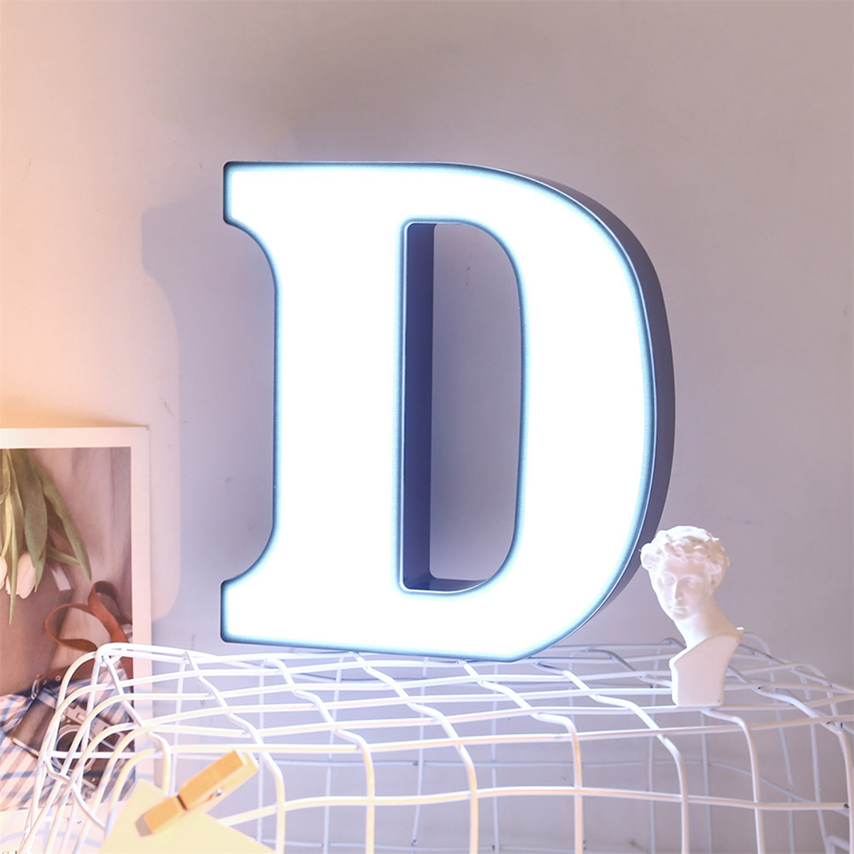 Ein großes, beleuchtetes 'D' an einer weißen Wand. Eine kleine Statue und ein gerahmtes Bild sind in der Nähe.