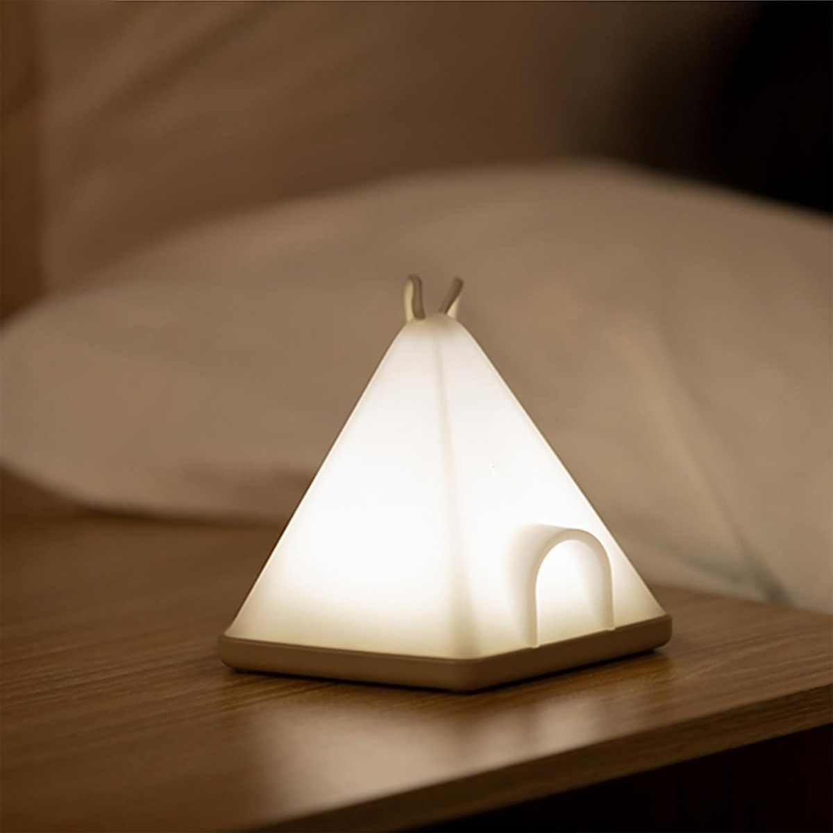 Eine leuchtende Tipi-Lampe auf einem hölzernen Nachttisch. Im Hintergrund ein weißes Kissen.